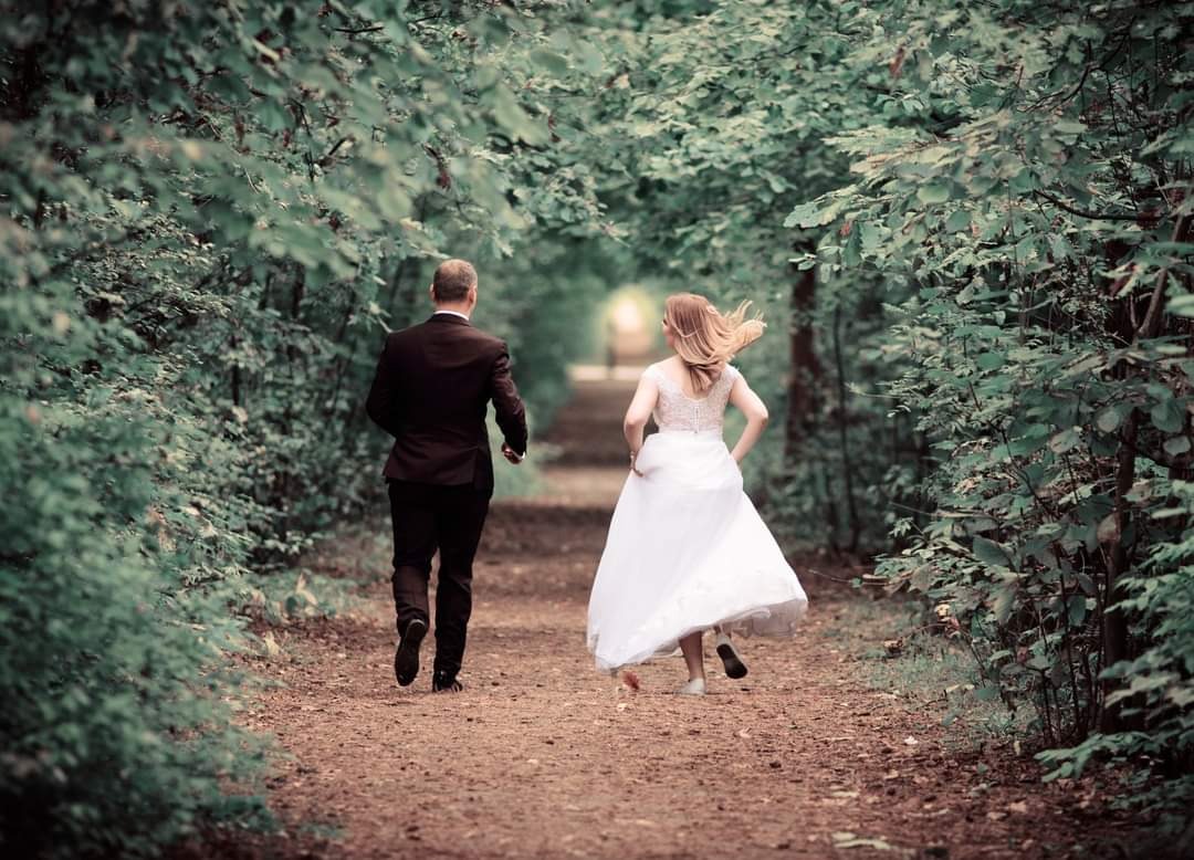Para młoda biegnie leśną ścieżką, panna młoda w białej sukni z koronkową górą, pan młody w ciemnym garniturze, widok z tyłu, tunel z zielonych drzew, światło na końcu ścieżki.