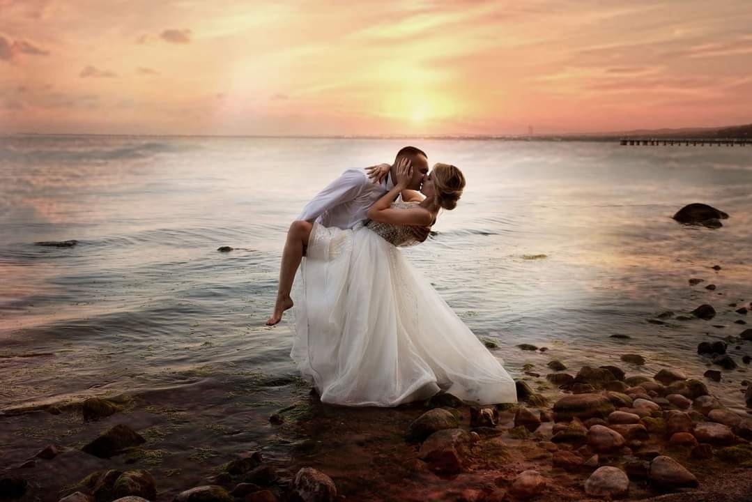 Para młoda w objęciach na brzegu morza o zachodzie słońca, panna młoda w białej sukni z odkrytymi ramionami, pan młody trzyma pannę młodą w ramionach, w tle woda i pomost