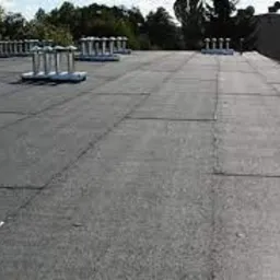 Płaski dach pokryty papą w arkuszach, widoczne kominki wentylacyjne i fragment ściany budynku w tle.