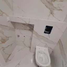 Wisząca toaleta w łazience wyłożonej białymi płytkami z marmurkowym wzorem, widoczna instalacja elektryczna i fragment zabudowy.