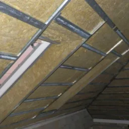 Poddasze w trakcie ocieplania wełną mineralną, widoczne metalowe profile i okno dachowe.