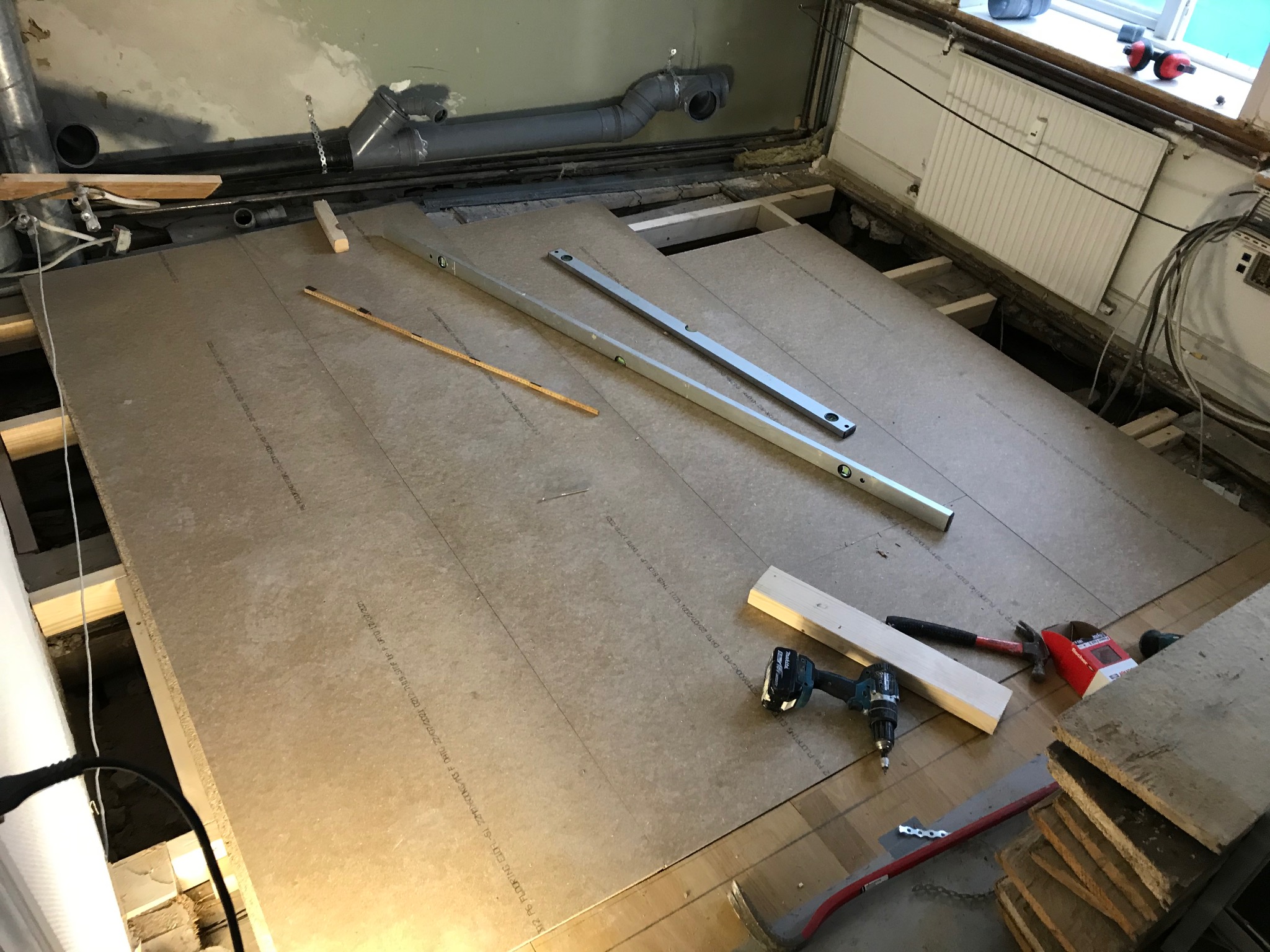 Montaż płyt podłogowych na legarach, widoczne poziomice, wkrętarka, młotek i paczka gwoździ, na tle instalacji wentylacyjnej pod sufitem i grzejnika przy oknie.