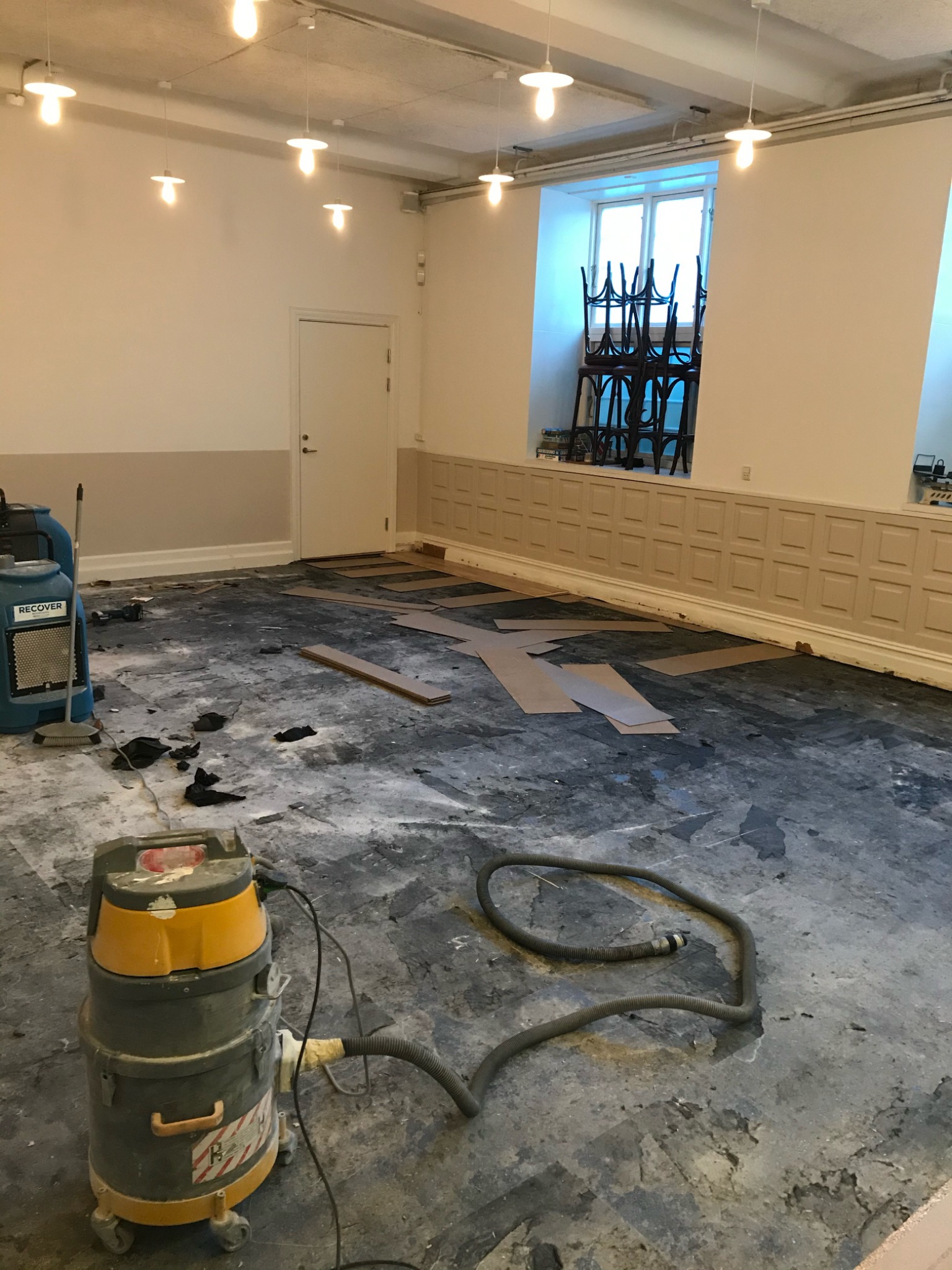 Przygotowanie podłoża pod parkiet w przestronnym pomieszczeniu z wysokimi oknami, widoczny odkurzacz przemysłowy, fragmenty paneli i maszyna do osuszania.