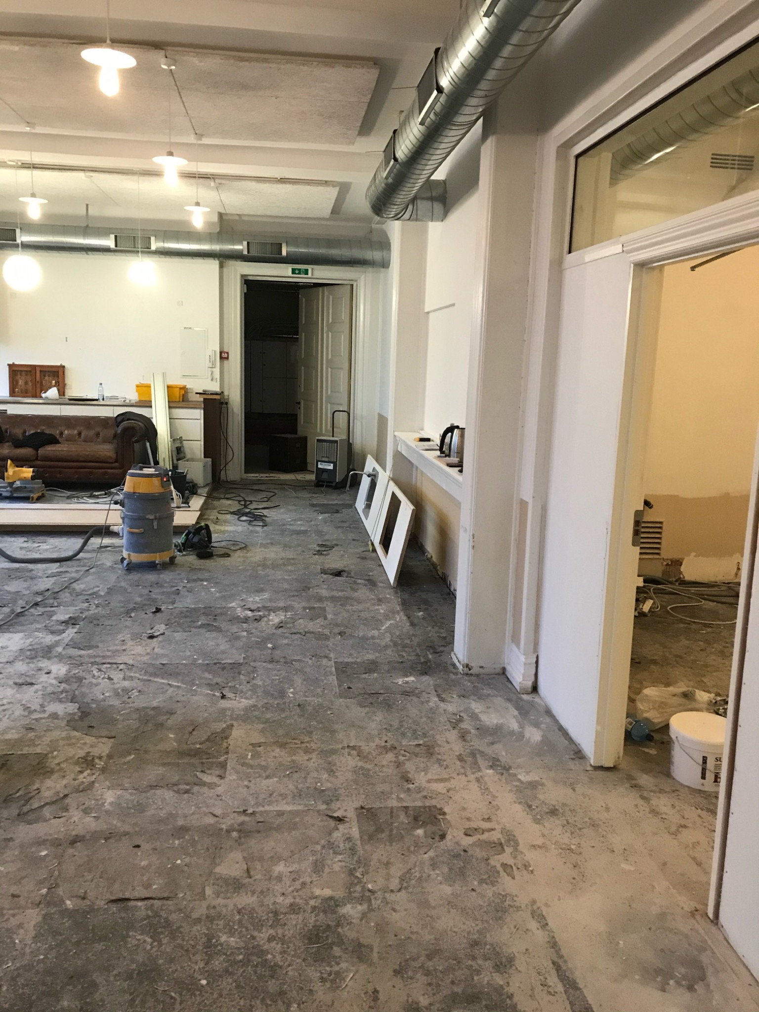 Wnętrze pomieszczenia w trakcie remontu: zerwane płytki podłogowe, odkurzacz przemysłowy, widoczne instalacje pod sufitem, przygotowania do dalszych prac.