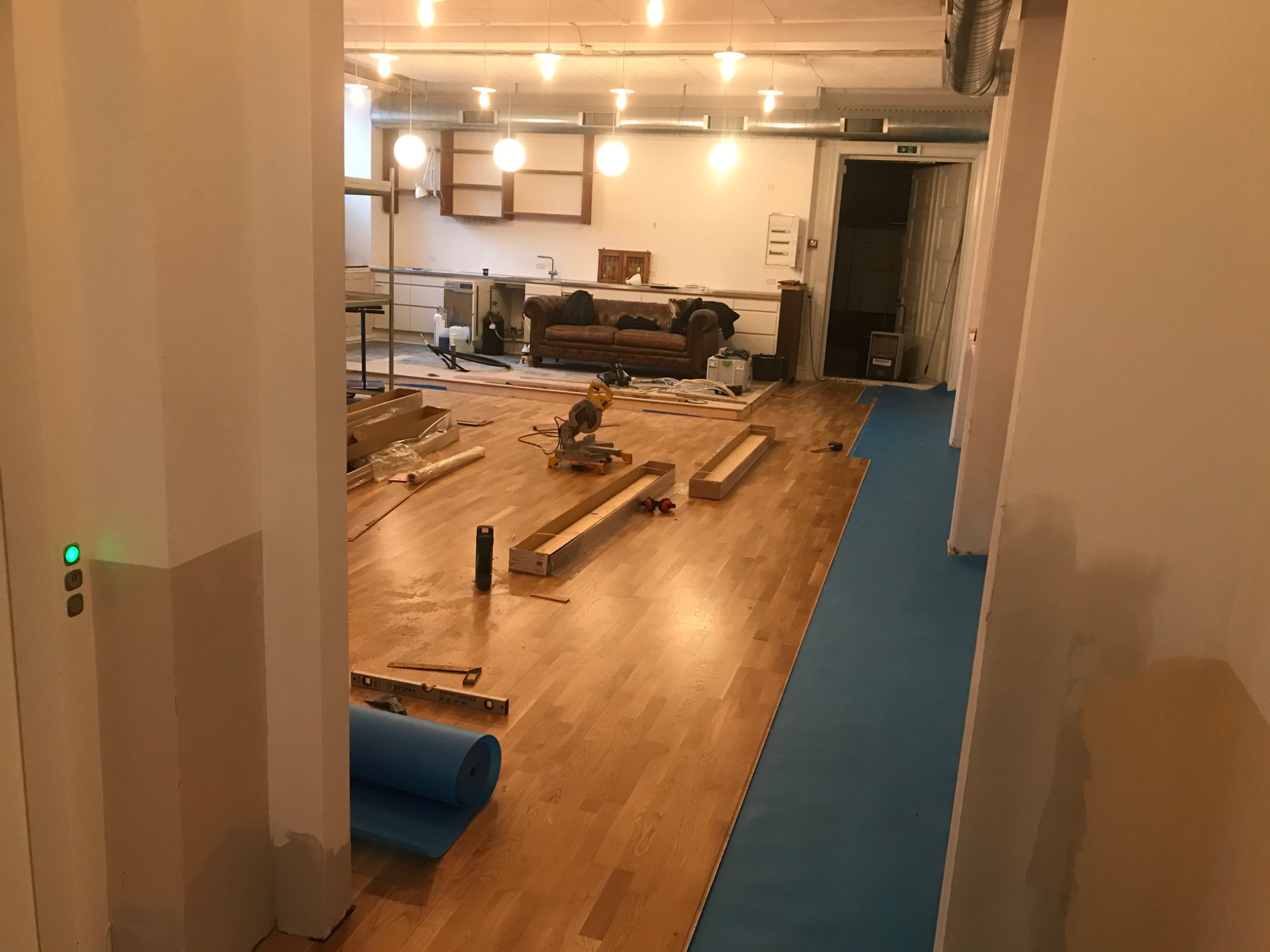 Parkiet dębowy układany w pomieszczeniu z aneksem kuchennym, widoczna maszyna do cięcia paneli i rolka niebieskiego podkładu wygłuszającego