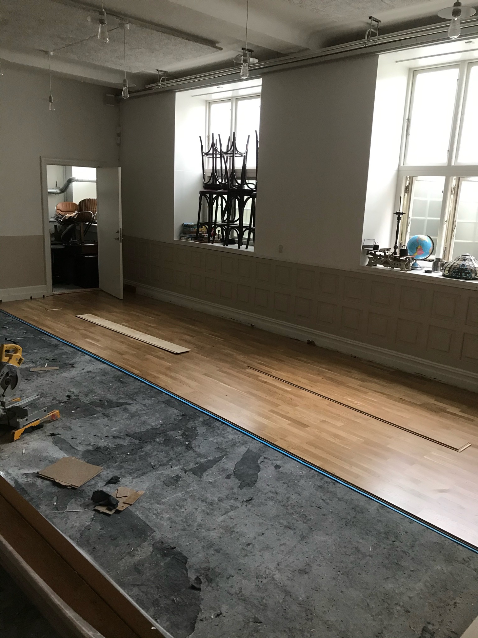 Układanie jasnego parkietu w przestronnym pomieszczeniu z wysokimi oknami, widoczne narzędzia do cięcia i fragmenty niezakończonej podłogi.