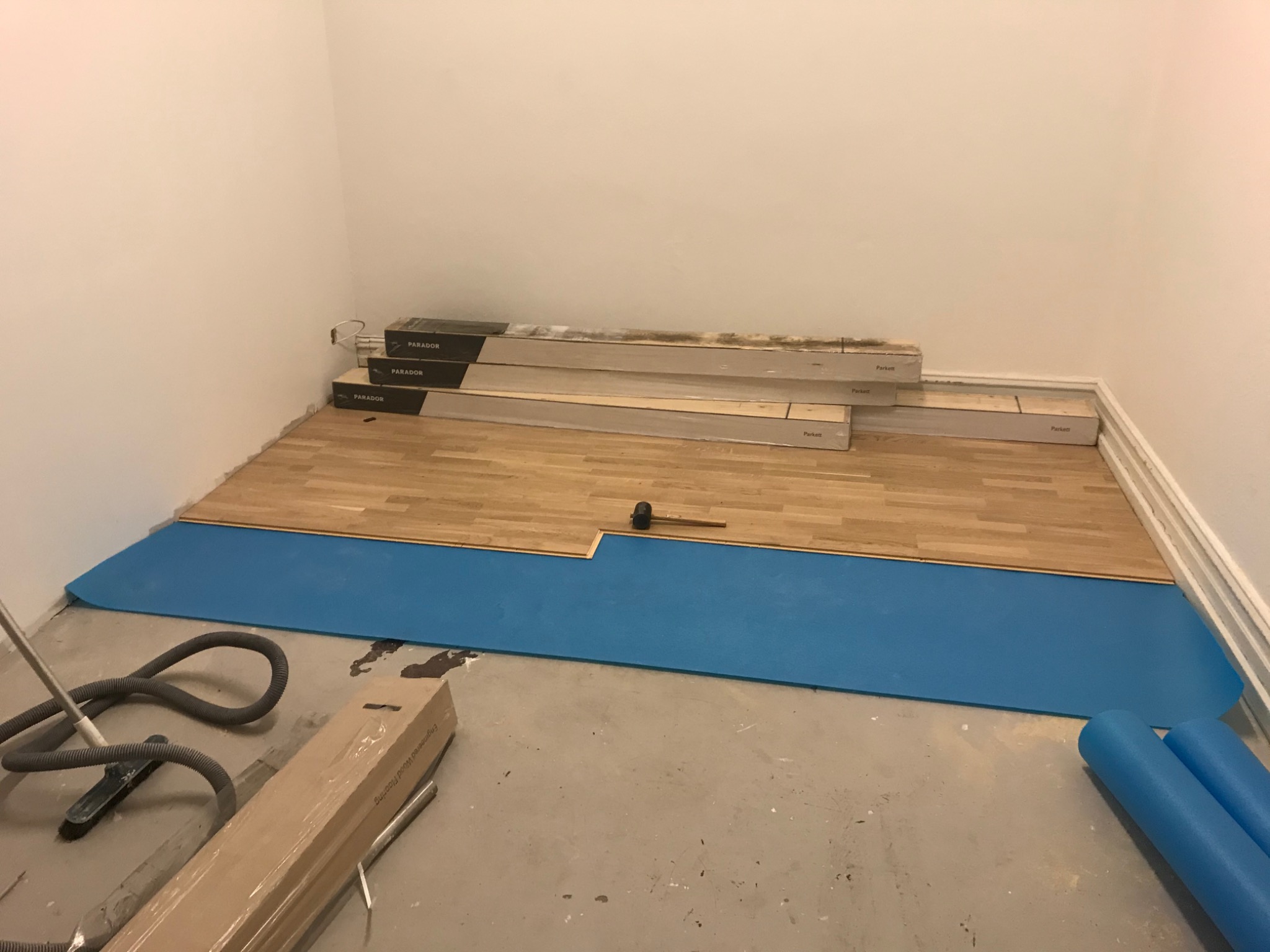 Proces układania drewnianego parkietu: częściowo ułożona podłoga, niebieski podkład, paczki paneli marki Parador przy ścianie, gumowy młotek i odkurzacz w tle.