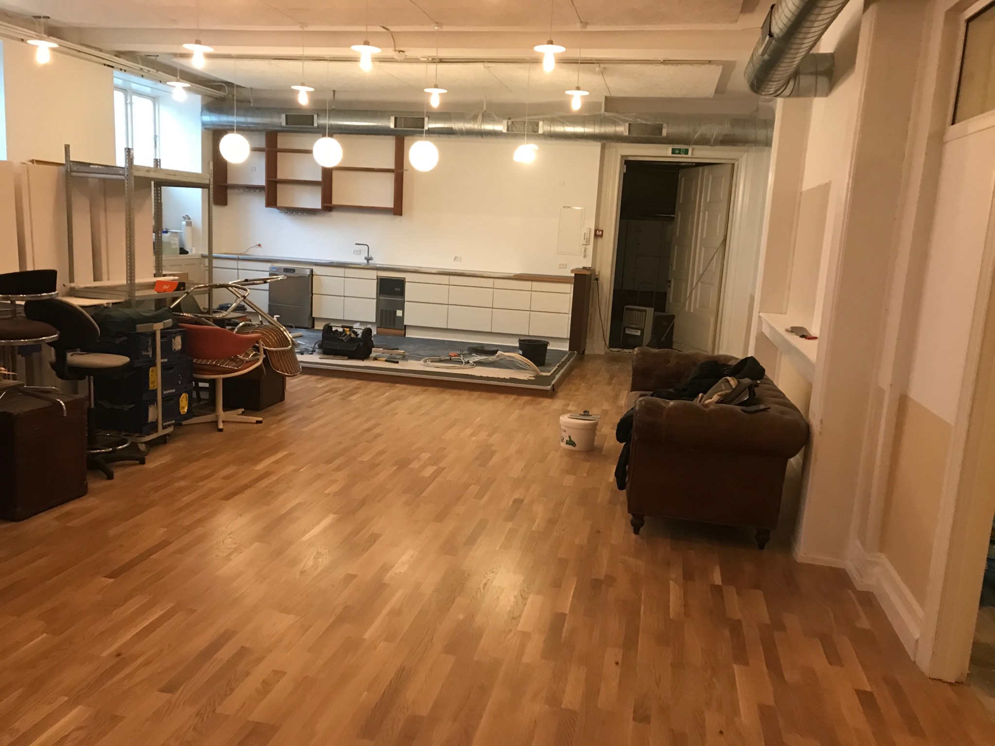 Przestronne wnętrze w trakcie prac: świeżo ułożony parkiet z widocznym rysunkiem drewna, częściowo wyposażona kuchnia z białymi szafkami, narzędzia i wiadro farby sugerują etap wykończeniowy.