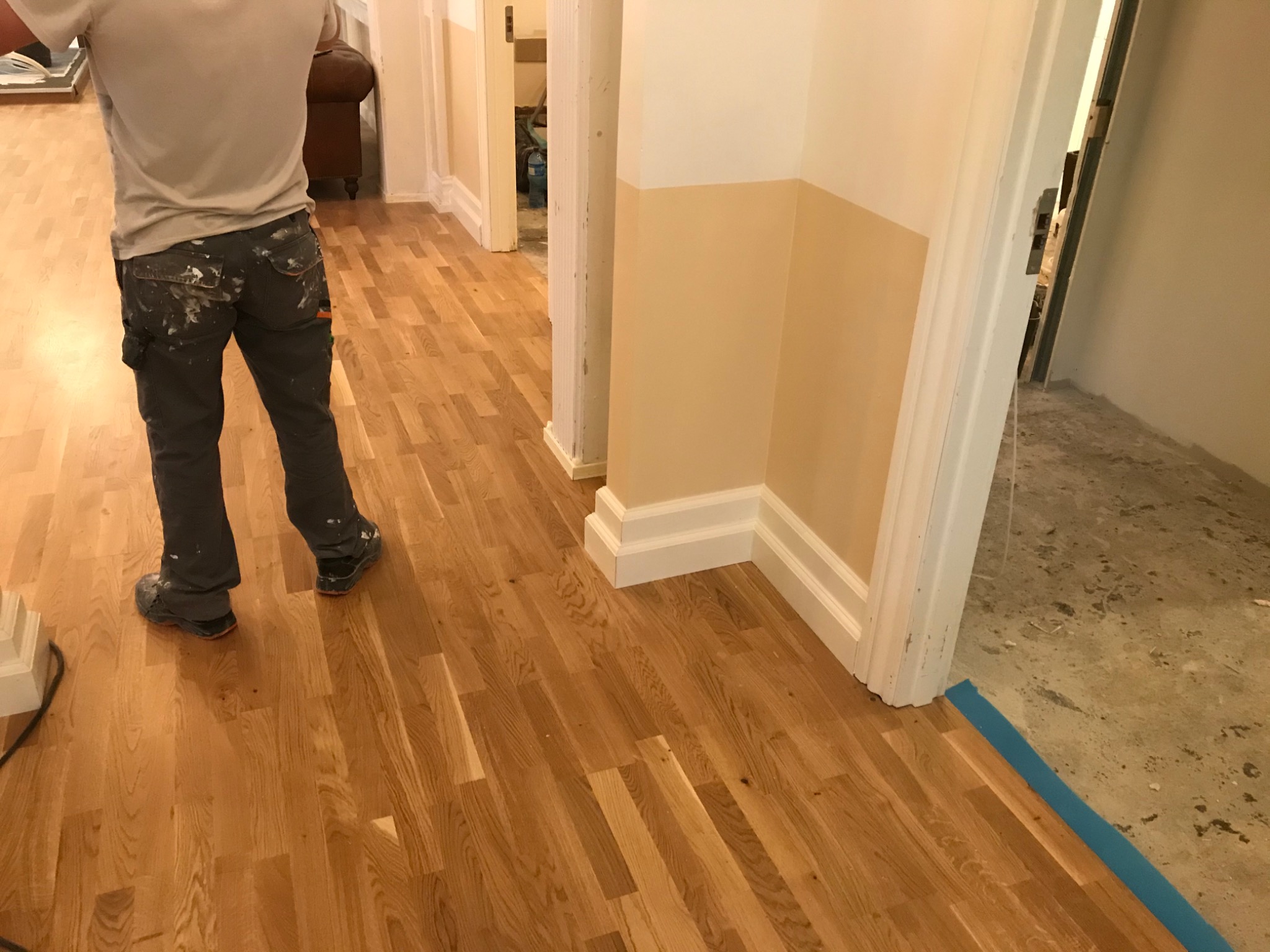 Parkiet ułożony w jodełkę, widok na korytarz w trakcie malowania, spodnie robocze malarza z plamami farby.