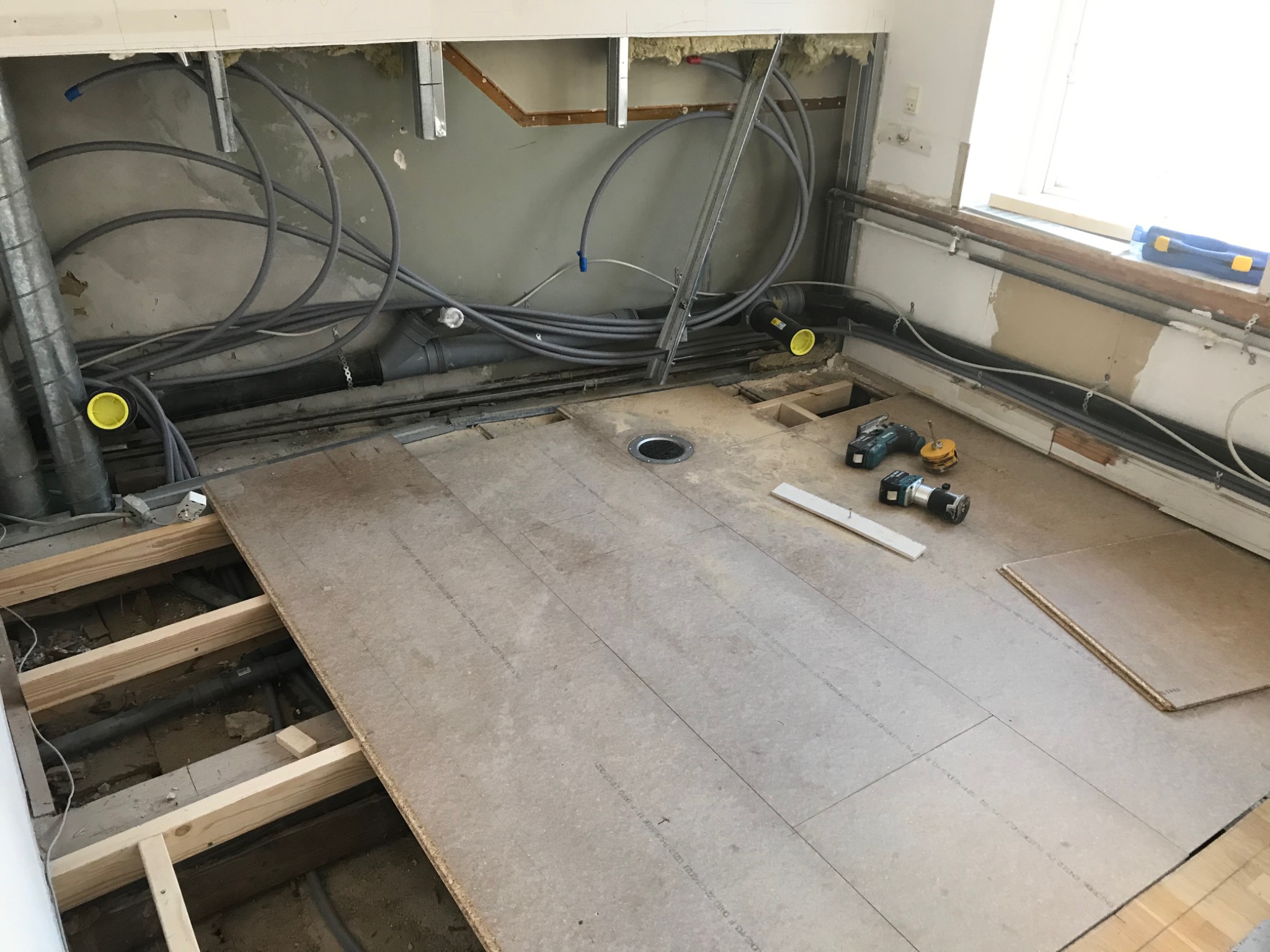 Płyta podłogowa ułożona na legarach, widoczne instalacje wodno-kanalizacyjne i elektryczne w ścianie, narzędzia pracy na podłodze, okno w tle.