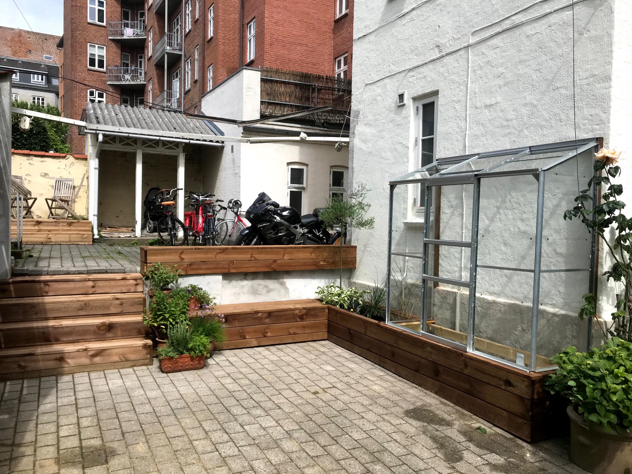 Ujęcie z poziomu gruntu na podwórko z kostki brukowej, drewnianymi schodami, podwyższonymi grządkami, małą szklarnią przy ścianie budynku, rowerami i motocyklem w tle.