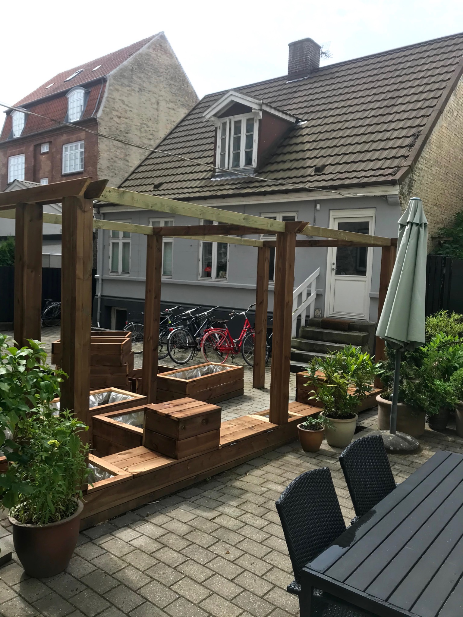 Drewniana konstrukcja pergoli z podwyższonymi grządkami w ogrodzie, widoczne donice z roślinami, rowery i budynki w tle.