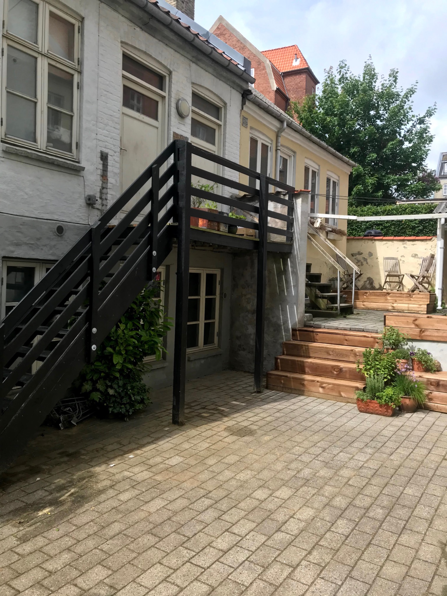 Dziedziniec kamienicy z drewnianymi schodami prowadzącymi na podest i do ogrodu, z roślinami w donicach i drewnianymi krzesłami w tle.