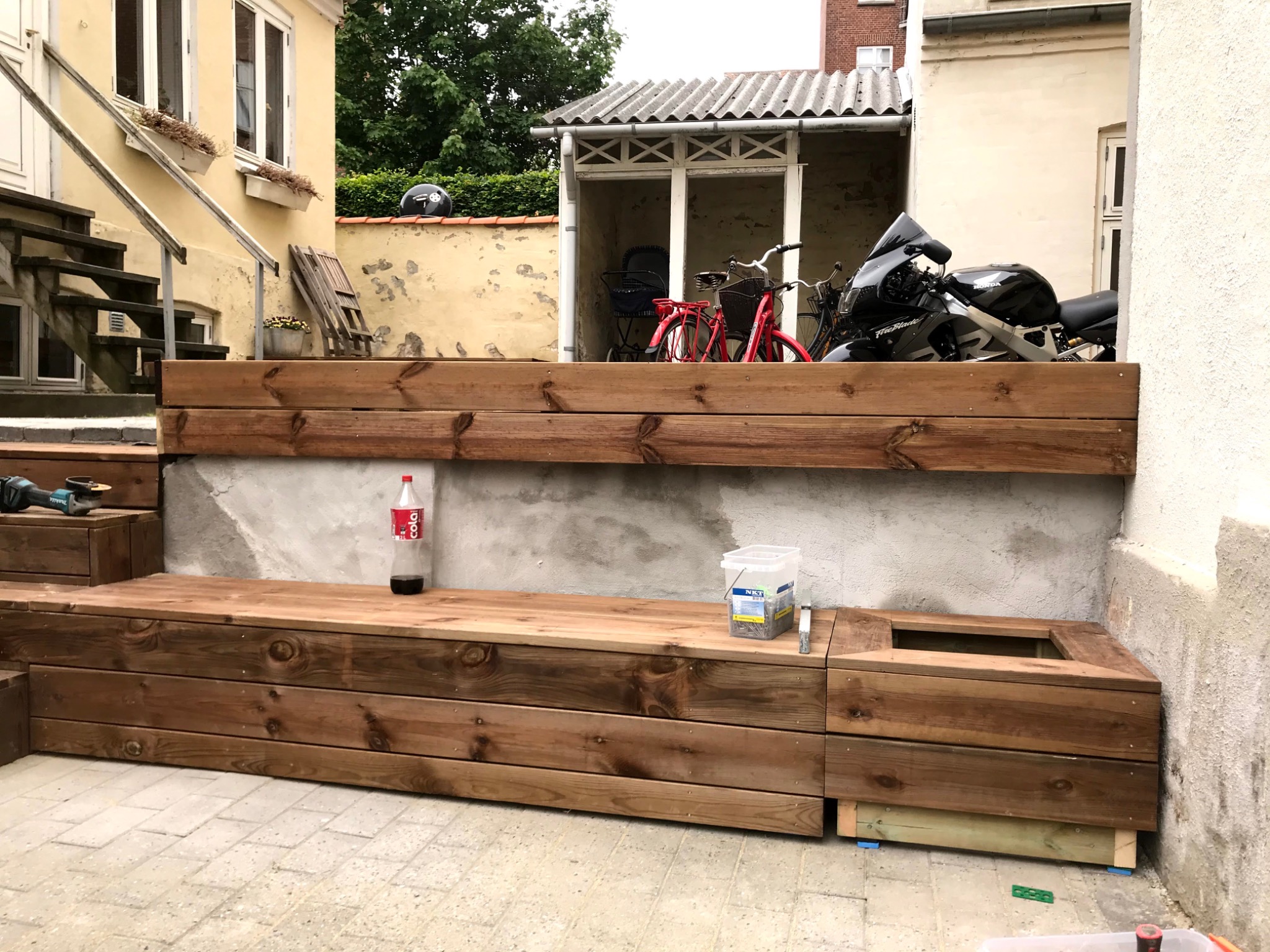 Drewniana konstrukcja tarasowa w trakcie budowy, z narzędziami i materiałami budowlanymi, butelką coli i widokiem na motocykl Honda Fireblade.