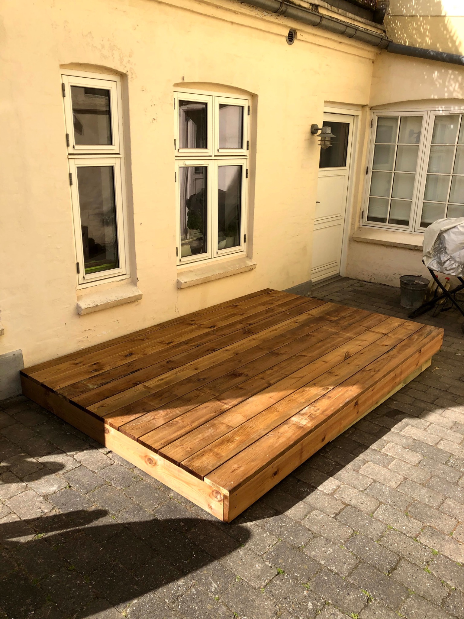 Drewniany taras z desek, z wyraźnymi słojami, przylegający do ściany budynku z oknami i drzwiami, ułożony na brukowanym podłożu.