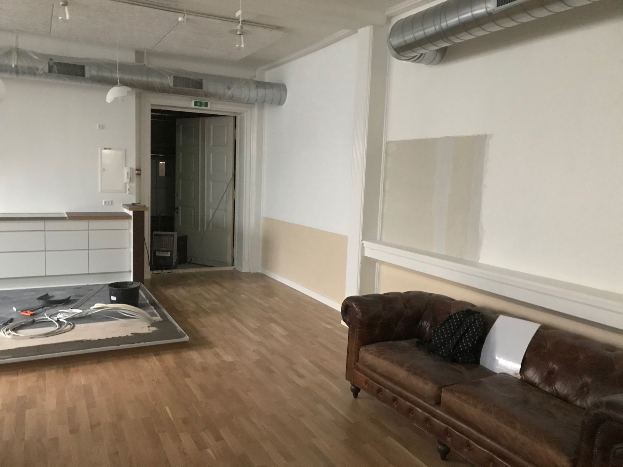 Przygotowanie do układania parkietu w pomieszczeniu z brązową skórzaną sofą, widoczne narzędzia i materiały na podkładzie, ściany częściowo pomalowane na biało i beżowo, wentylacja pod sufitem.