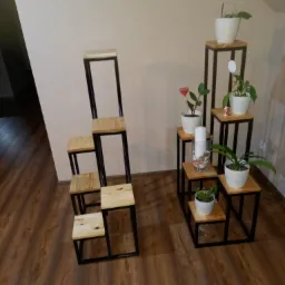 Dwie metalowo-drewniane konstrukcje schodkowe, pełniące funkcję kwietników, z roślinami w białych doniczkach, ustawione na brązowej podłodze w pomieszczeniu.