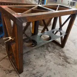 Stalowa konstrukcja stołu z kratownicą i półką z otworami, surowy stan z widocznymi śladami spawania i rdzawym nalotem, uchwyt spawalniczy przypięty z boku.