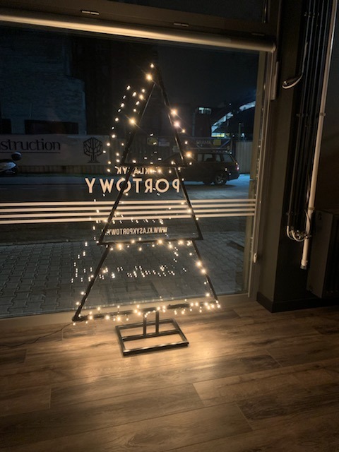 Metalowa konstrukcja w kształcie choinki, oświetlona małymi lampkami, widoczna przez szybę z napisem reklamowym, na tle nocnego krajobrazu miejskiego.