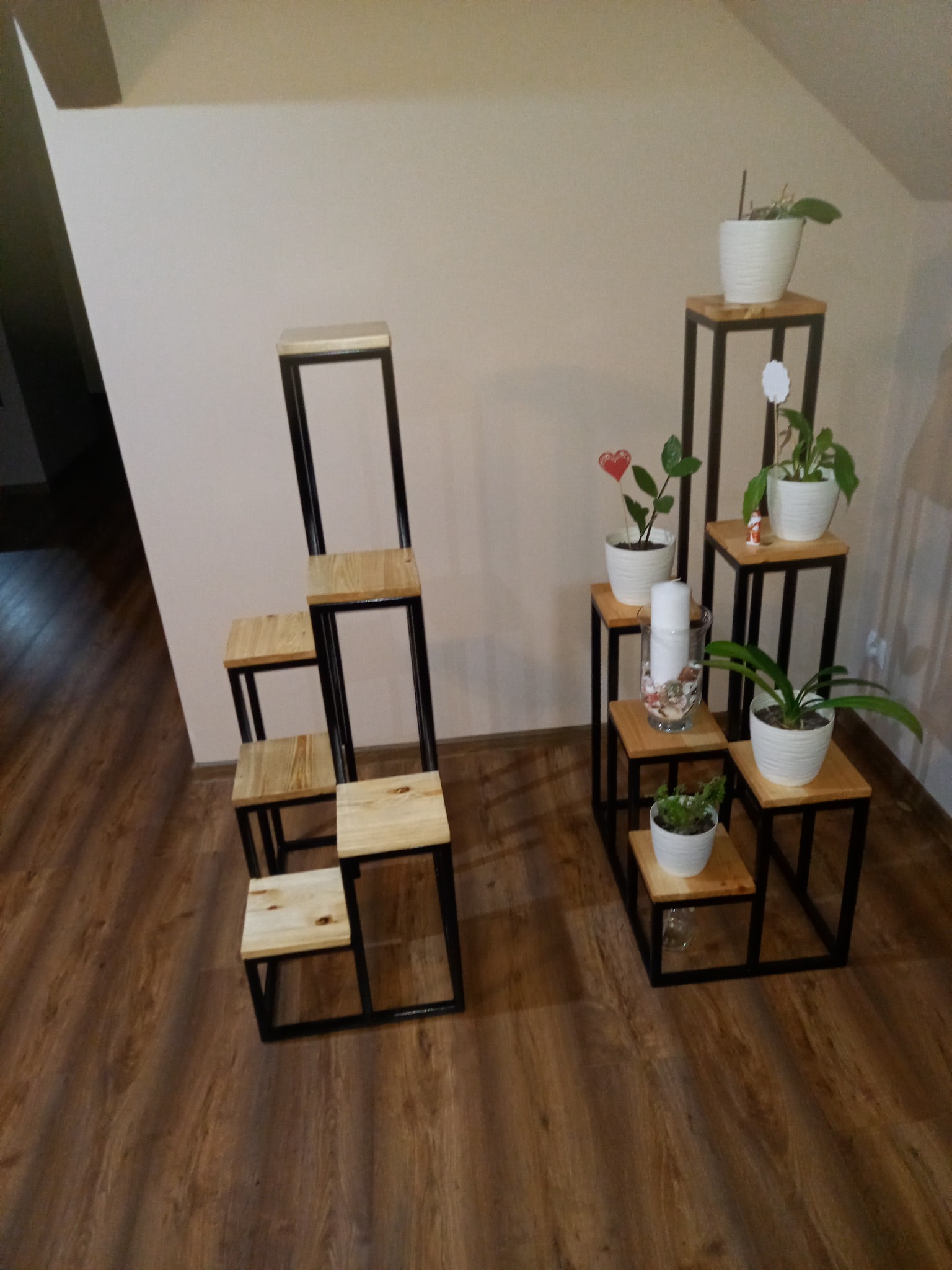 Dwie metalowo-drewniane konstrukcje schodkowe, pełniące funkcję kwietników, z roślinami w białych doniczkach, ustawione na brązowej podłodze w pomieszczeniu.