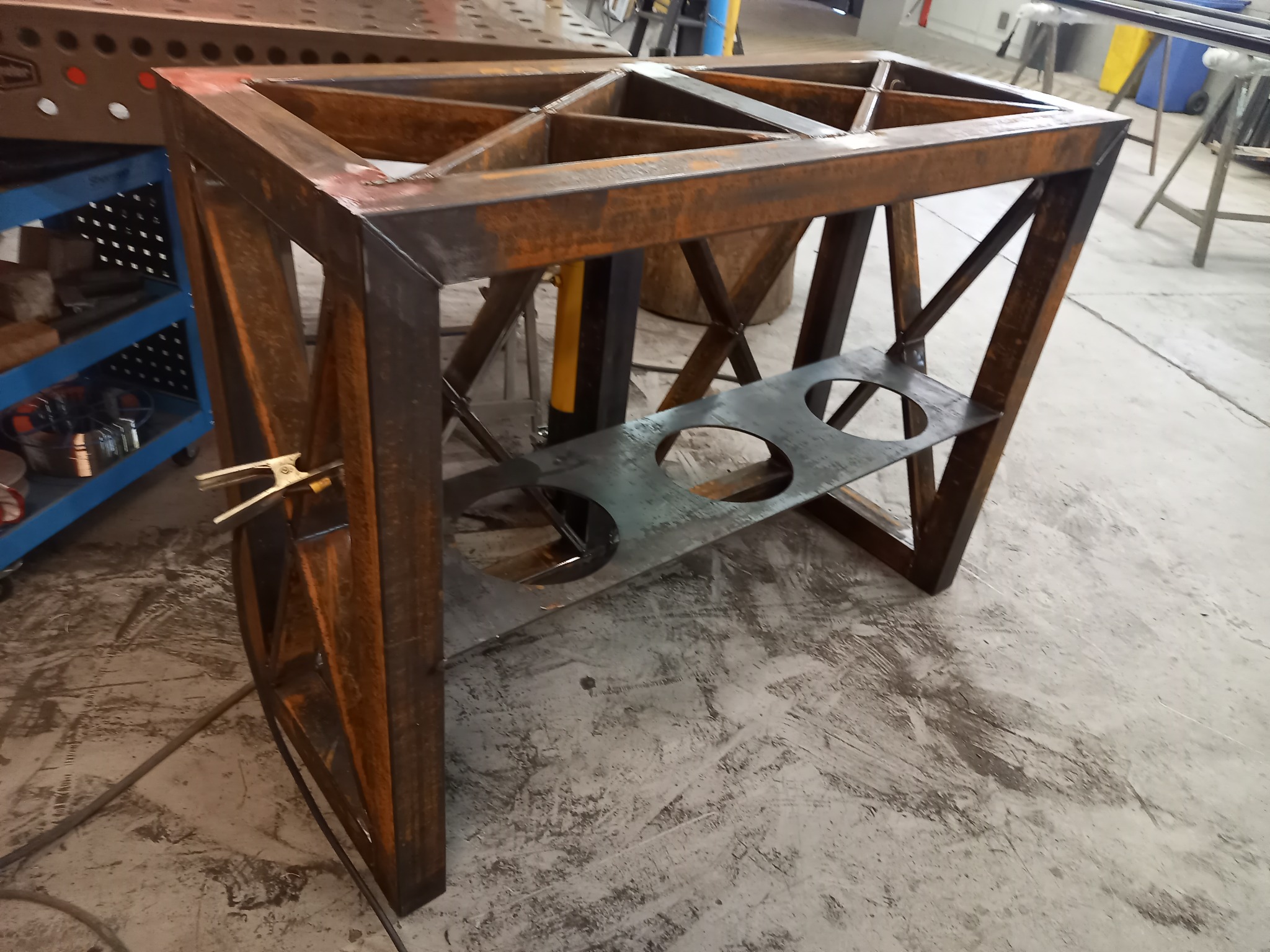 Stalowa konstrukcja stołu z kratownicą i półką z otworami, surowy stan z widocznymi śladami spawania i rdzawym nalotem, uchwyt spawalniczy przypięty z boku.