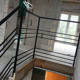 LOFT STYL usługi ślusarsko-spawalnicze - Surowe wnętrze w trakcie budowy z widoczną czarną, metalową balustradą schodową o minimalistycznym designie, zamontowaną przy ścianie z betonu komórkowego, częściowo wykończoną, z widocznymi...
