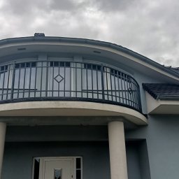 LOFT STYL usługi ślusarsko-spawalnicze - Półokrągły balkon z czarną, metalową balustradą z motywem rombu, wsparty na dwóch kolumnach, na tle szarej elewacji domu i pochmurnego nieba.