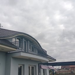 LOFT STYL usługi ślusarsko-spawalnicze - Szary dom jednorodzinny z balkonem zabezpieczonym czarną metalową balustradą, widok z dołu na tle pochmurnego nieba.