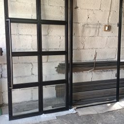 LOFT STYL usługi ślusarsko-spawalnicze - Częściowo zmontowane, czarne metalowe drzwi z kwadratowymi szybami z siatki, opierające się o białą ceglaną ścianę w warsztacie, obok ułożone metalowe profile.