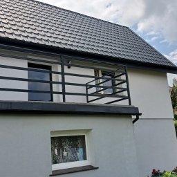 LOFT STYL usługi ślusarsko-spawalnicze - Nowoczesny balkon z czarną, metalową balustradą o poziomym układzie prętów, zamontowany przy białym domu z ciemną dachówką, widok z ogrodu w słoneczny dzień.