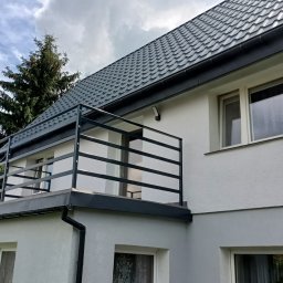 LOFT STYL usługi ślusarsko-spawalnicze - Nowoczesna, metalowa balustrada balkonowa w Złotoryi, z poziomymi szczeblami, zamontowana na balkonie domu z szarą dachówką. Widok z dołu, na tle błękitnego nieba z chmurami.