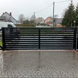 LOFT STYL usługi ślusarsko-spawalnicze - Nowoczesna, pozioma brama wjazdowa w kolorze czarnym, z widocznym mechanizmem otwierania, na tle mokrej kostki brukowej i z lekko widocznym samochodem dostawczym.