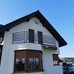 LOFT STYL usługi ślusarsko-spawalnicze - Dwupiętrowy dom z jasną elewacją, ciemnym dachem, balkonem z metalową balustradą i brązowymi roletami zewnętrznymi, sfotografowany w słoneczny dzień z niskiej perspektywy.
