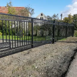 Czarna metalowa brama wjazdowa z dekoracyjnym wzorem meandrycznym i motywem krzyża, na tle domu z czerwoną dachówką i błękitnego nieba.