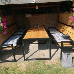 LOFT STYL usługi ślusarsko-spawalnicze - Zadaszona altana z drewnianym stołem i metalowymi ławkami z poduchami, ozdobiona wiszącymi kwiatami, ustawiona na kamiennej posadzce.