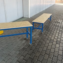 LOFT STYL usługi ślusarsko-spawalnicze - Dwa metalowe stoły warsztatowe z drewnianymi blatami z płyty OSB na kółkach, ustawione na zewnątrz budynku o białej fasadzie i kostce brukowej.
