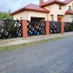 LOFT STYL usługi ślusarsko-spawalnicze - Czarny, metalowy płot o geometrycznym wzorze, z ceglanymi słupkami, na tle beżowego domu z czerwoną dachówką i otwartym garażem.