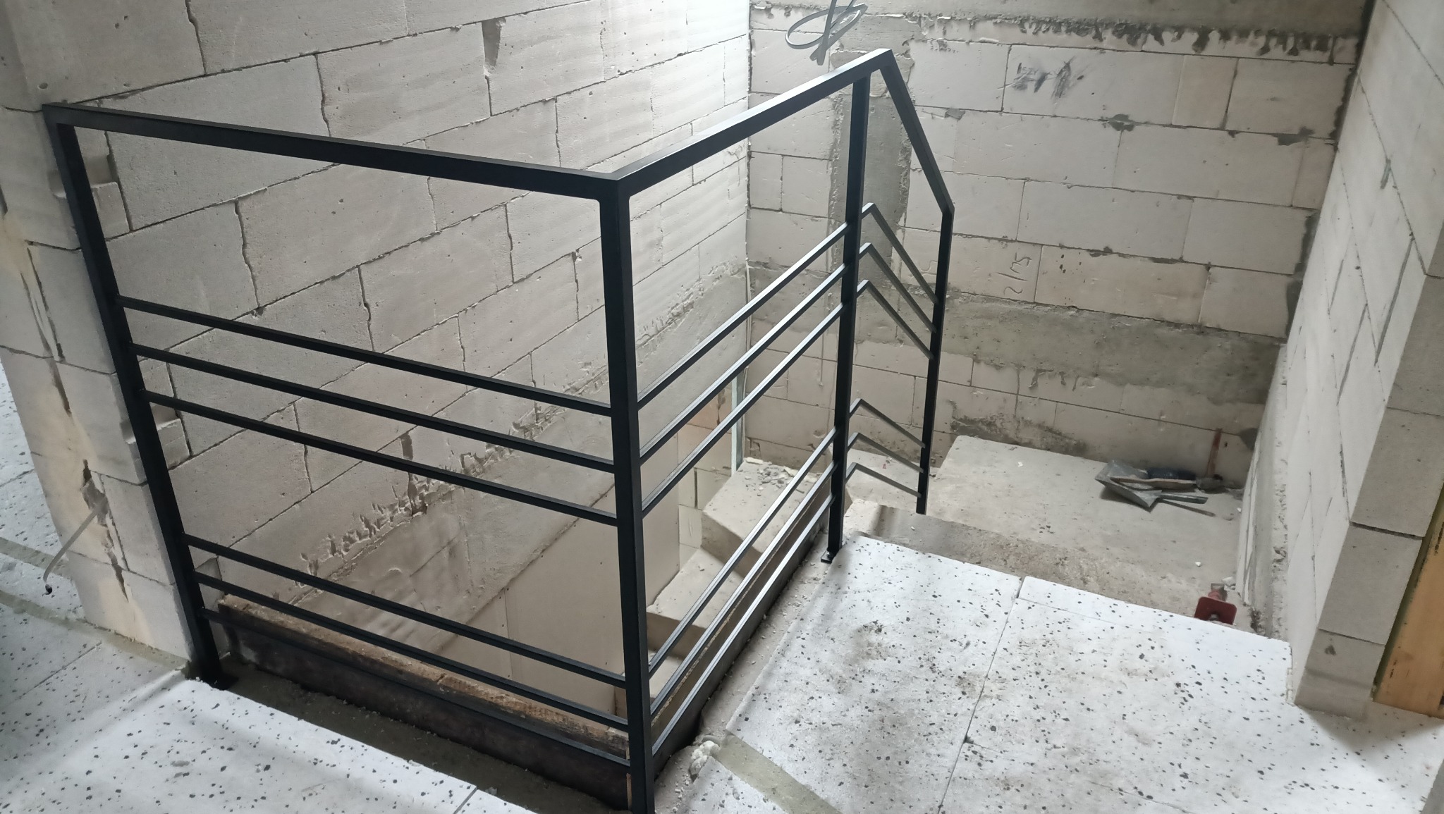 Metalowa balustrada schodowa z poziomymi prętami, zamontowana w surowym wnętrzu z bloczków betonowych i narzędziami budowlanymi w tle.