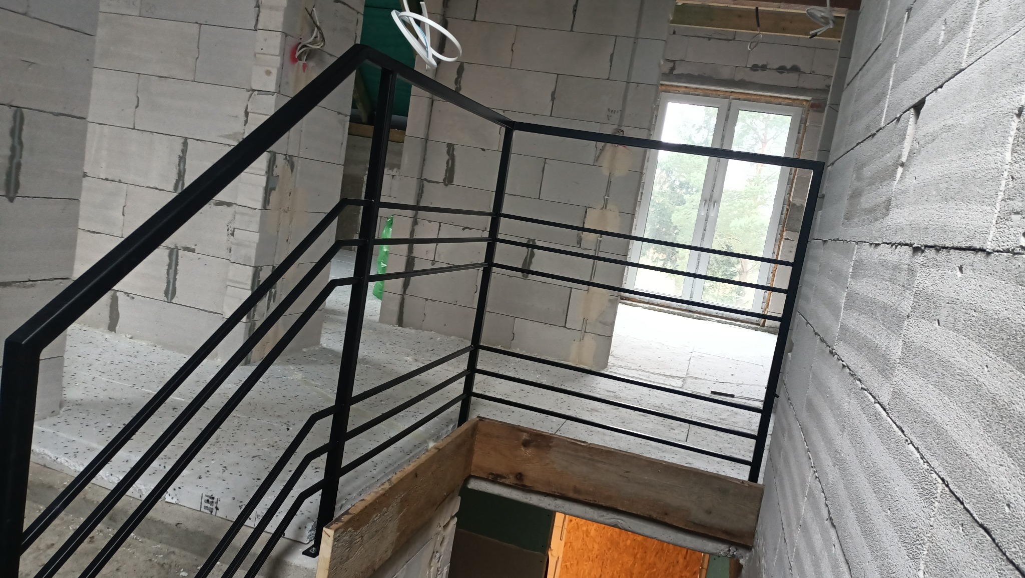 Surowe wnętrze w trakcie budowy z widoczną czarną, metalową balustradą schodową o minimalistycznym designie, zamontowaną przy ścianie z betonu komórkowego, częściowo wykończoną, z widocznymi...