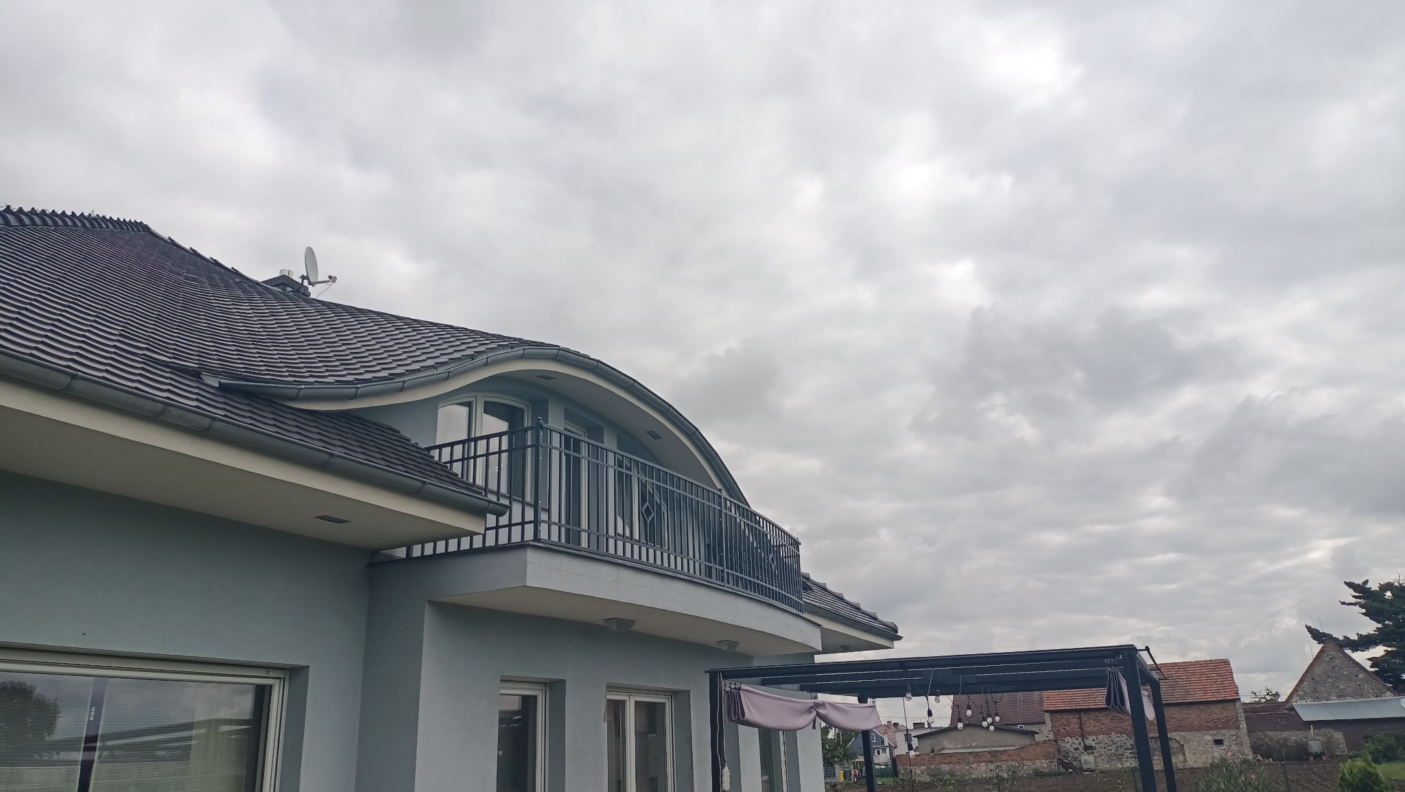 Szary dom jednorodzinny z balkonem zabezpieczonym czarną metalową balustradą, widok z dołu na tle pochmurnego nieba.