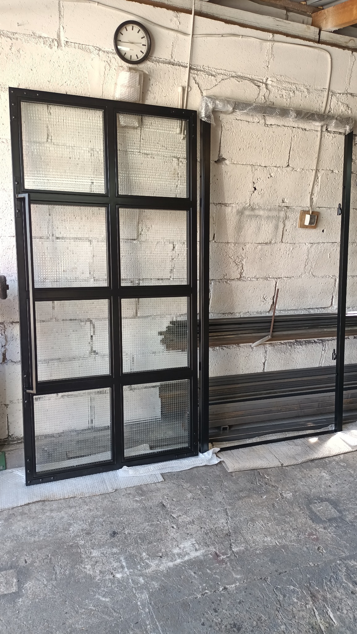 Częściowo zmontowane, czarne metalowe drzwi z kwadratowymi szybami z siatki, opierające się o białą ceglaną ścianę w warsztacie, obok ułożone metalowe profile.