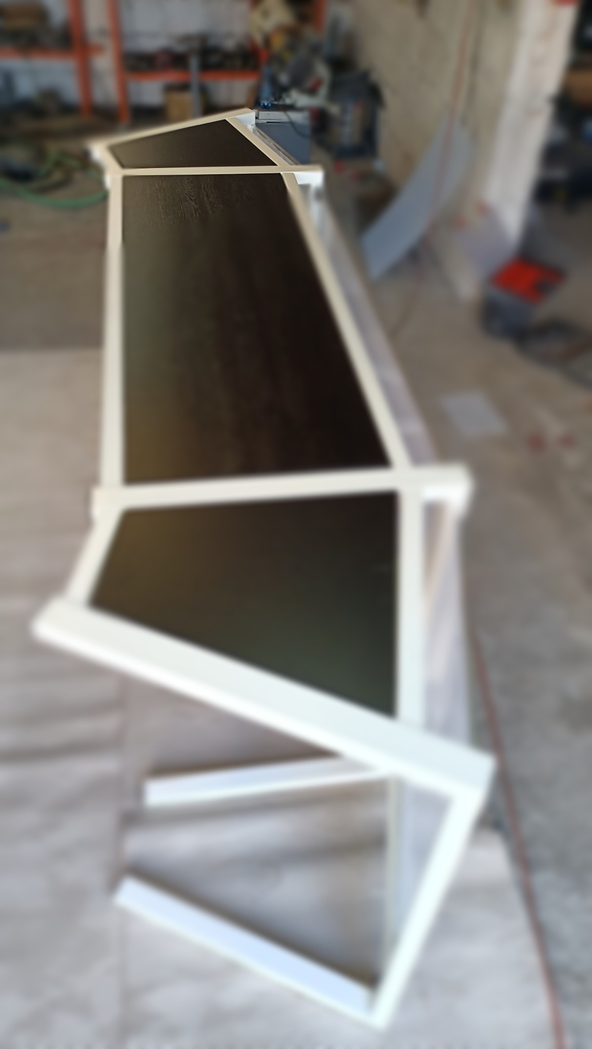 Metalowa konstrukcja w trakcie budowy, z ciemnymi panelami i białymi profilami, widziana pod kątem w warsztacie.