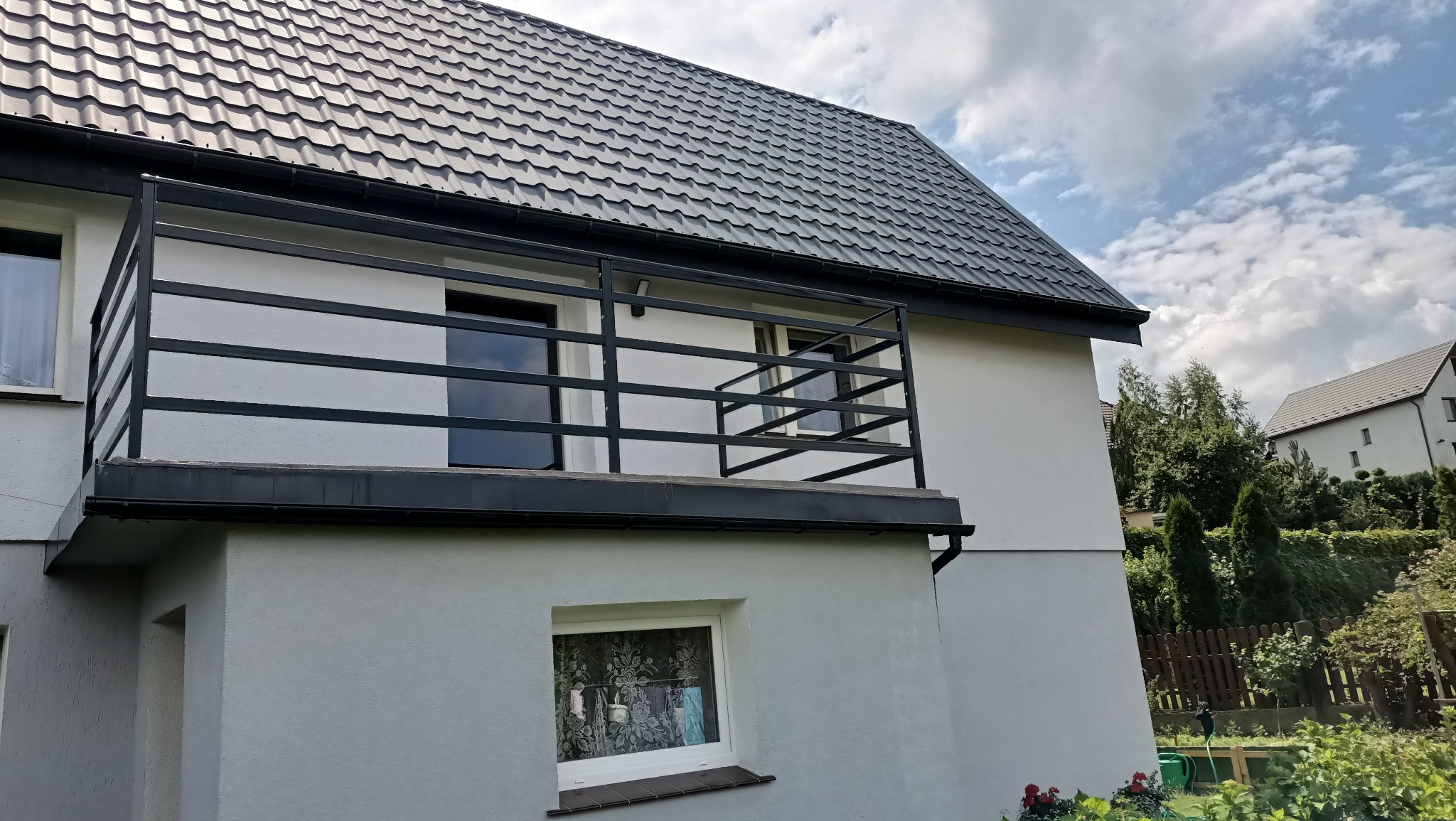 Nowoczesny balkon z czarną, metalową balustradą o poziomym układzie prętów, zamontowany przy białym domu z ciemną dachówką, widok z ogrodu w słoneczny dzień.