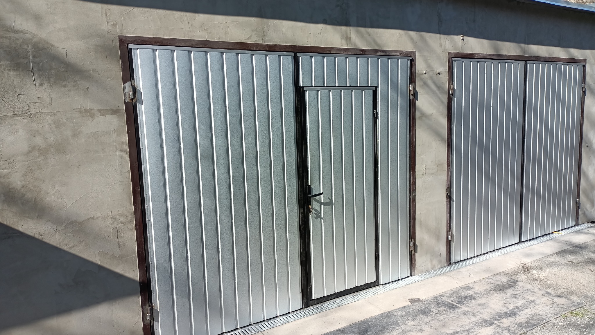 Dwa segmentowe, blaszane wrota garażowe z brązowymi ramami, jedno z dodatkowymi drzwiczkami, zamontowane w szarym, surowym murze.