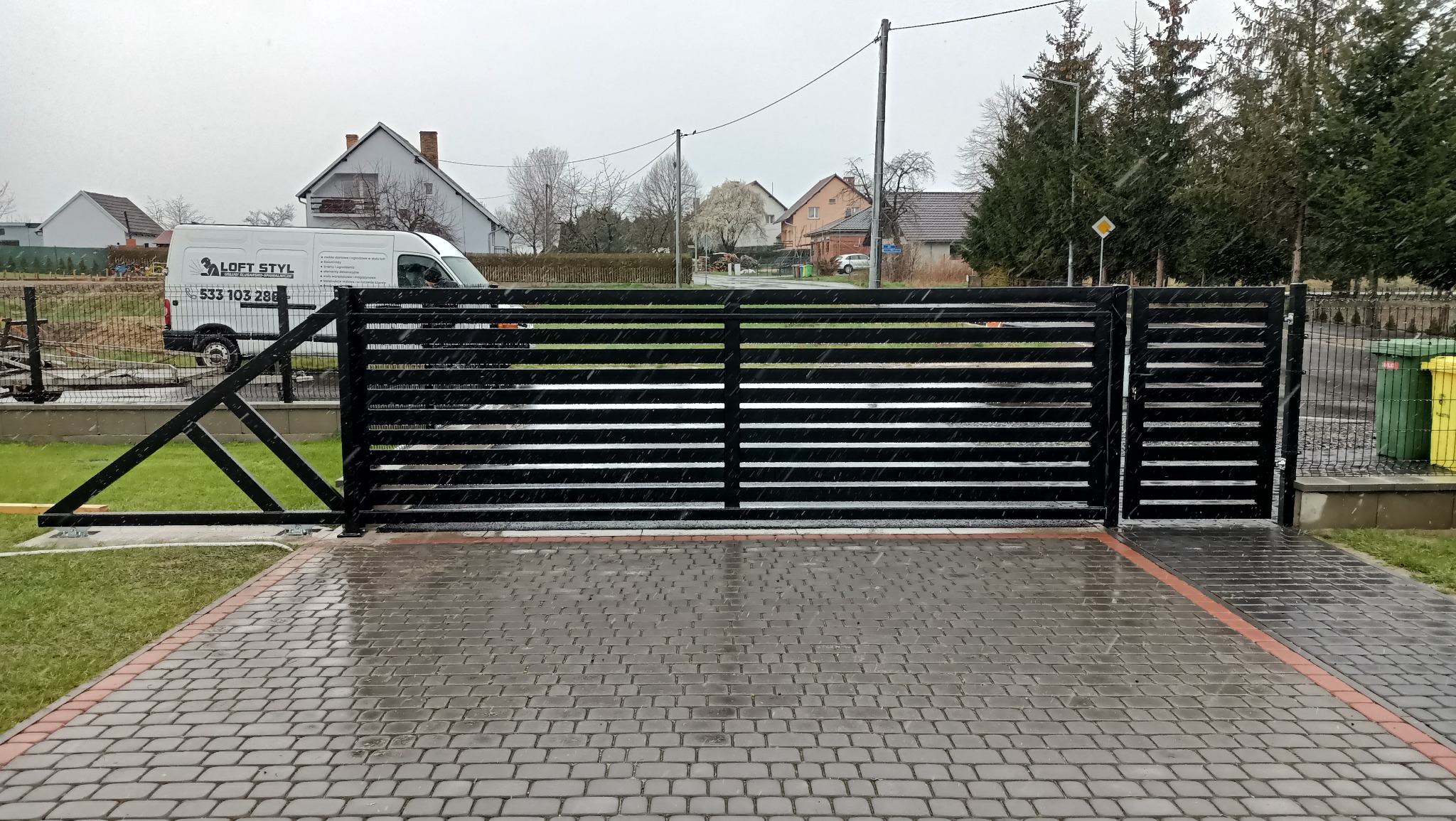 Nowoczesna, pozioma brama wjazdowa w kolorze czarnym, z widocznym mechanizmem otwierania, na tle mokrej kostki brukowej i z lekko widocznym samochodem dostawczym.