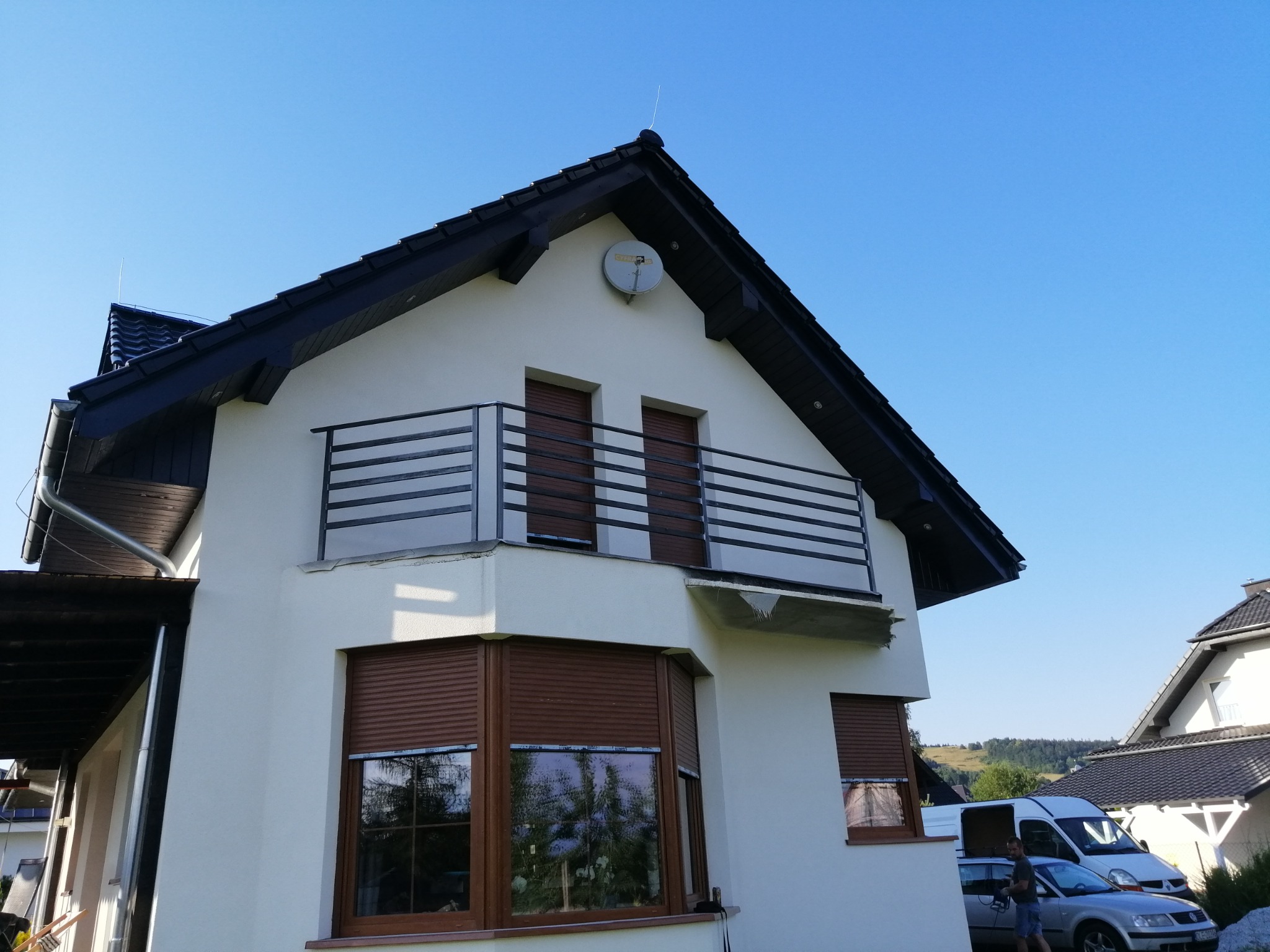 Dwupiętrowy dom z jasną elewacją, ciemnym dachem, balkonem z metalową balustradą i brązowymi roletami zewnętrznymi, sfotografowany w słoneczny dzień z niskiej perspektywy.