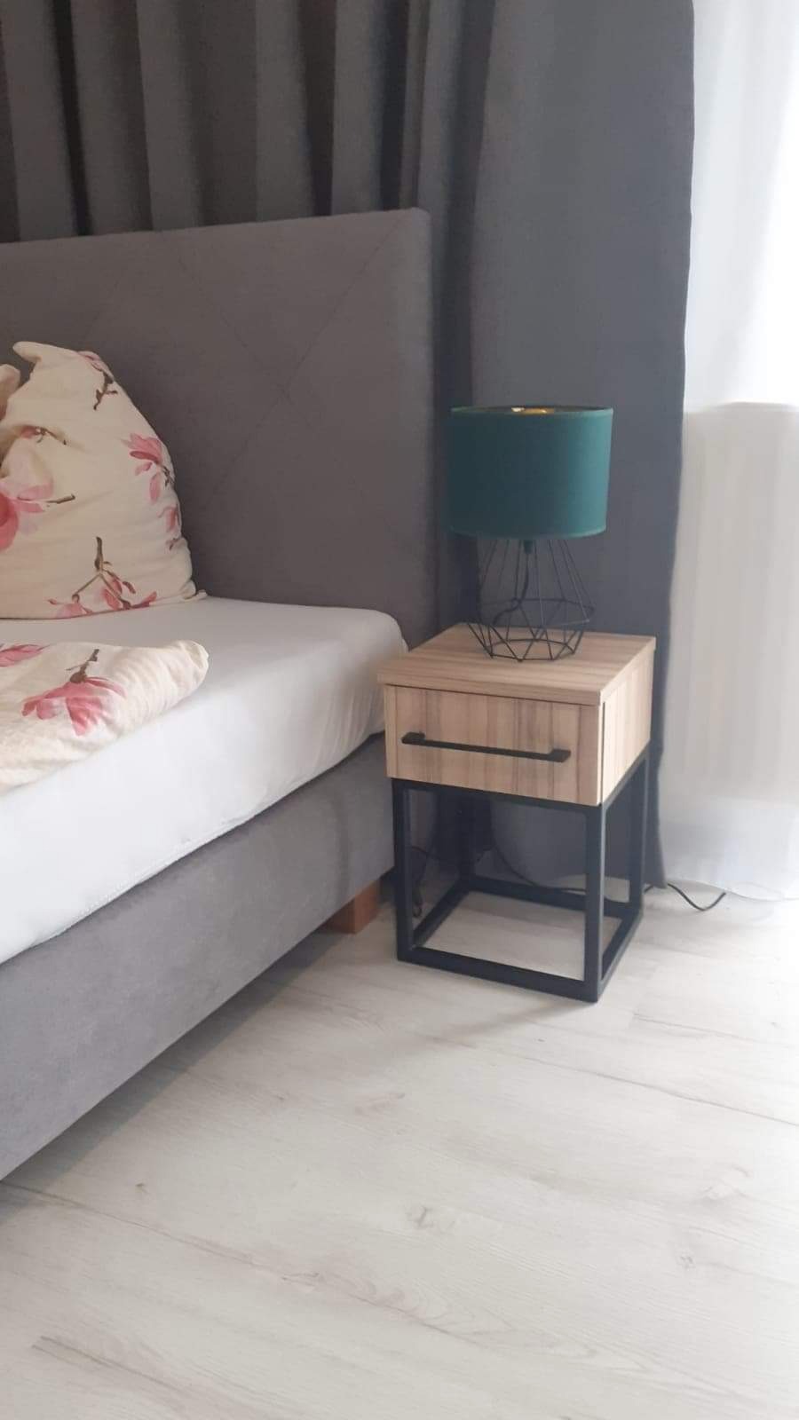 Nowoczesna szafka nocna z drewnianym frontem i czarną metalową ramą, na której stoi zielona lampka nocna z geometrycznym abażurem, obok łóżka z szarą tapicerką i poduszką w kwiaty.