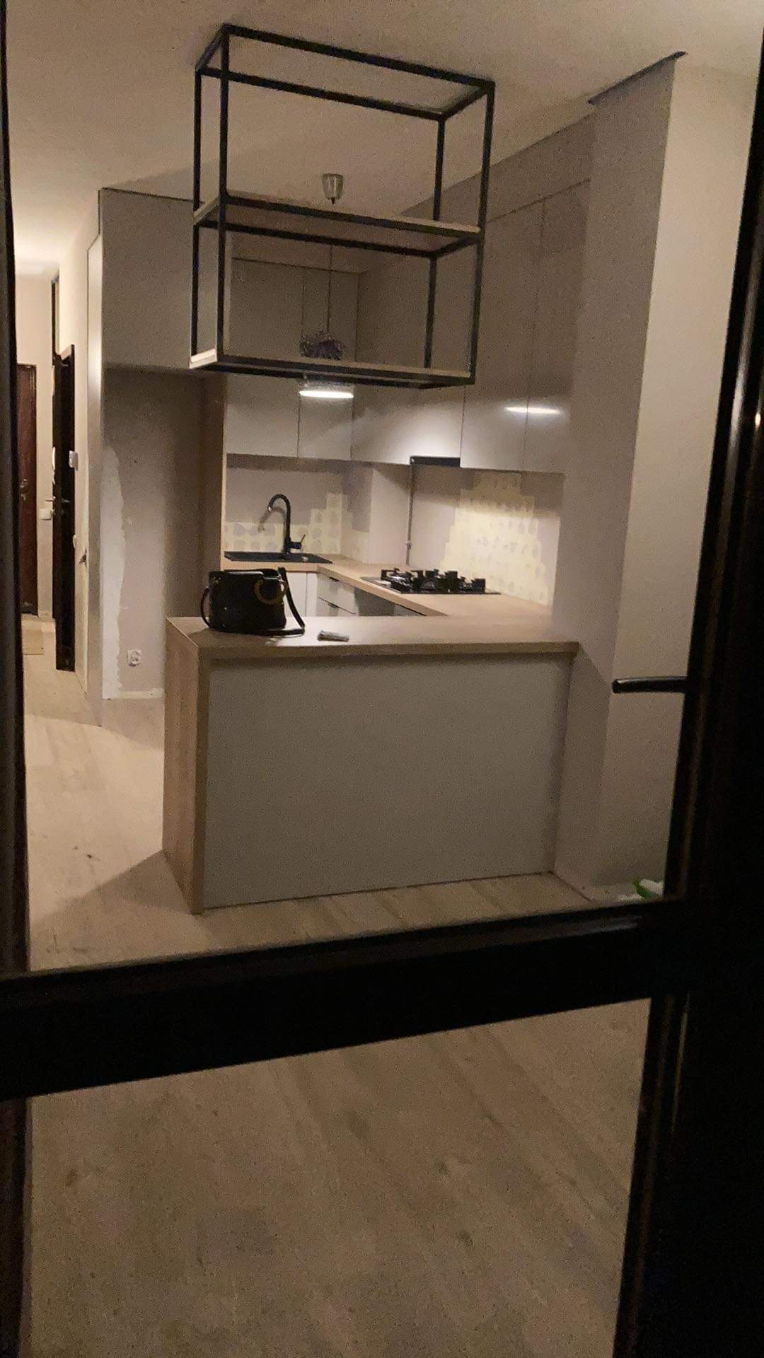 Wnętrze kuchni w trakcie montażu, widok przez framugę drzwi, wyspa kuchenna z drewnianym blatem, czarna armatura, płyta grzewcza, na blacie torba i narzędzie, nad wyspą metalowa konstrukcja sufitowa.