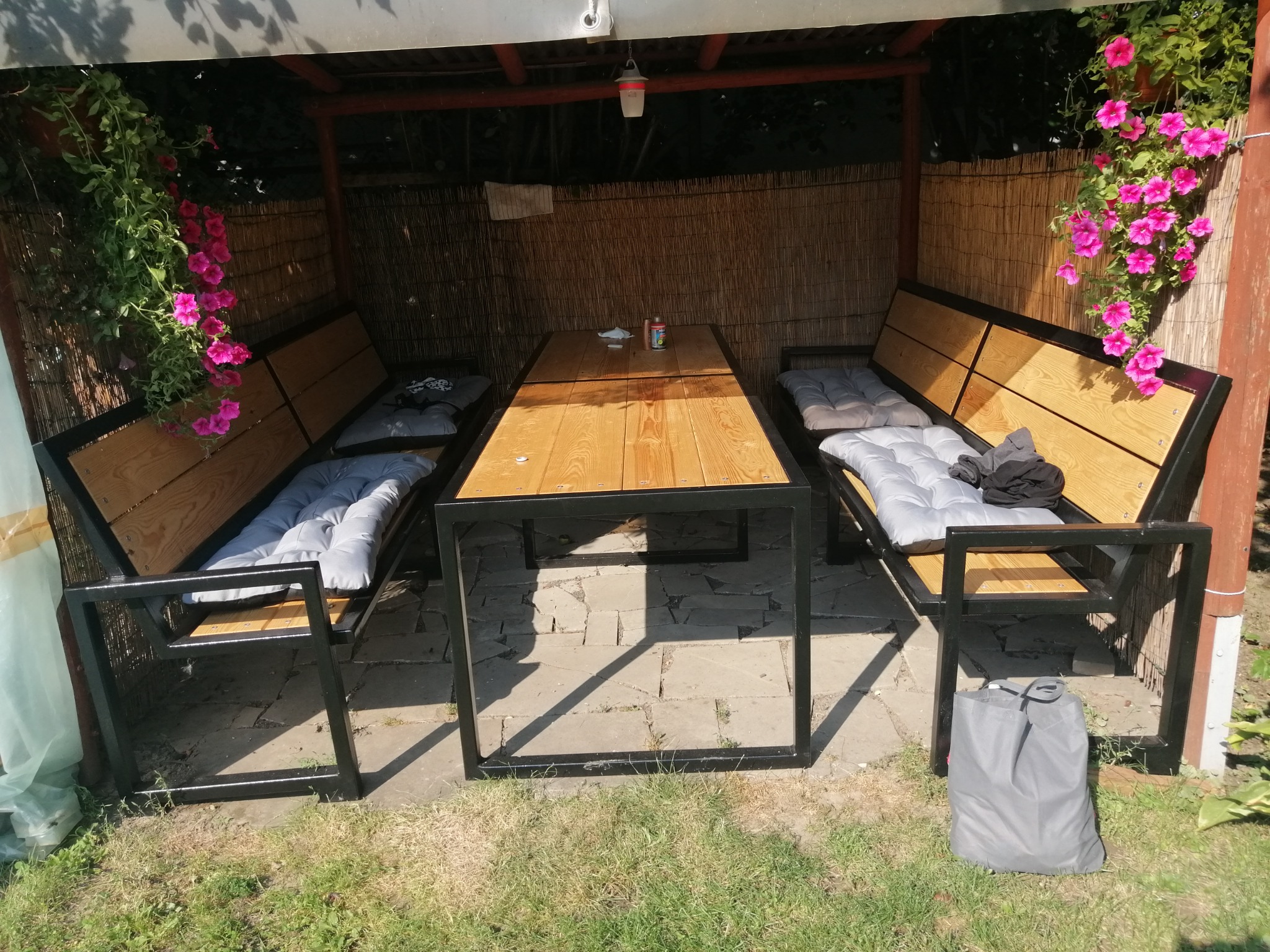 Zadaszona altana z drewnianym stołem i metalowymi ławkami z poduchami, ozdobiona wiszącymi kwiatami, ustawiona na kamiennej posadzce.