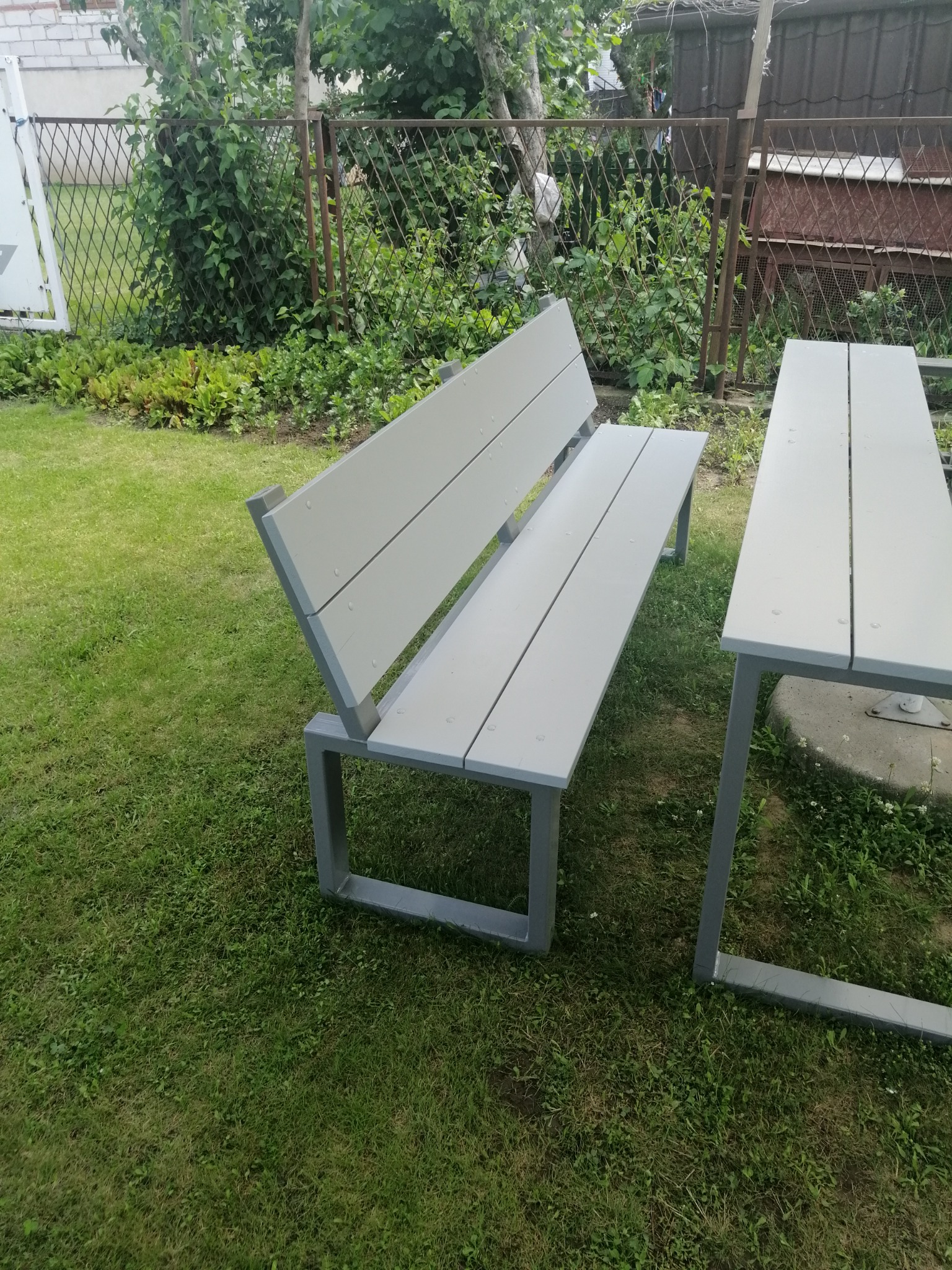 Szary metalowy stół i ławka ogrodowa na trawniku, widok z boku, w tle siatka ogrodzeniowa i zieleń.